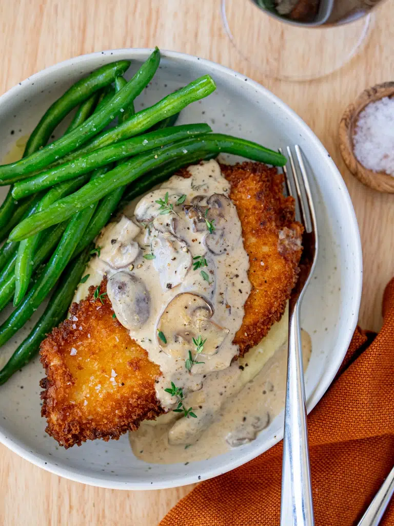 Crispy breaded chicken schnitzel topped with creamy mushroom sauce, served with sautéed green beans and mashed potatoes in a bowl.