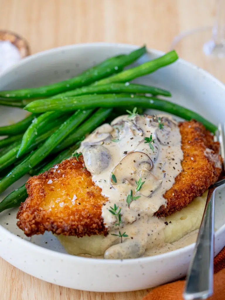 Golden crispy schnitzel topped with creamy mushroom sauce, served over mashed potatoes with a side of green beans in a bowl.