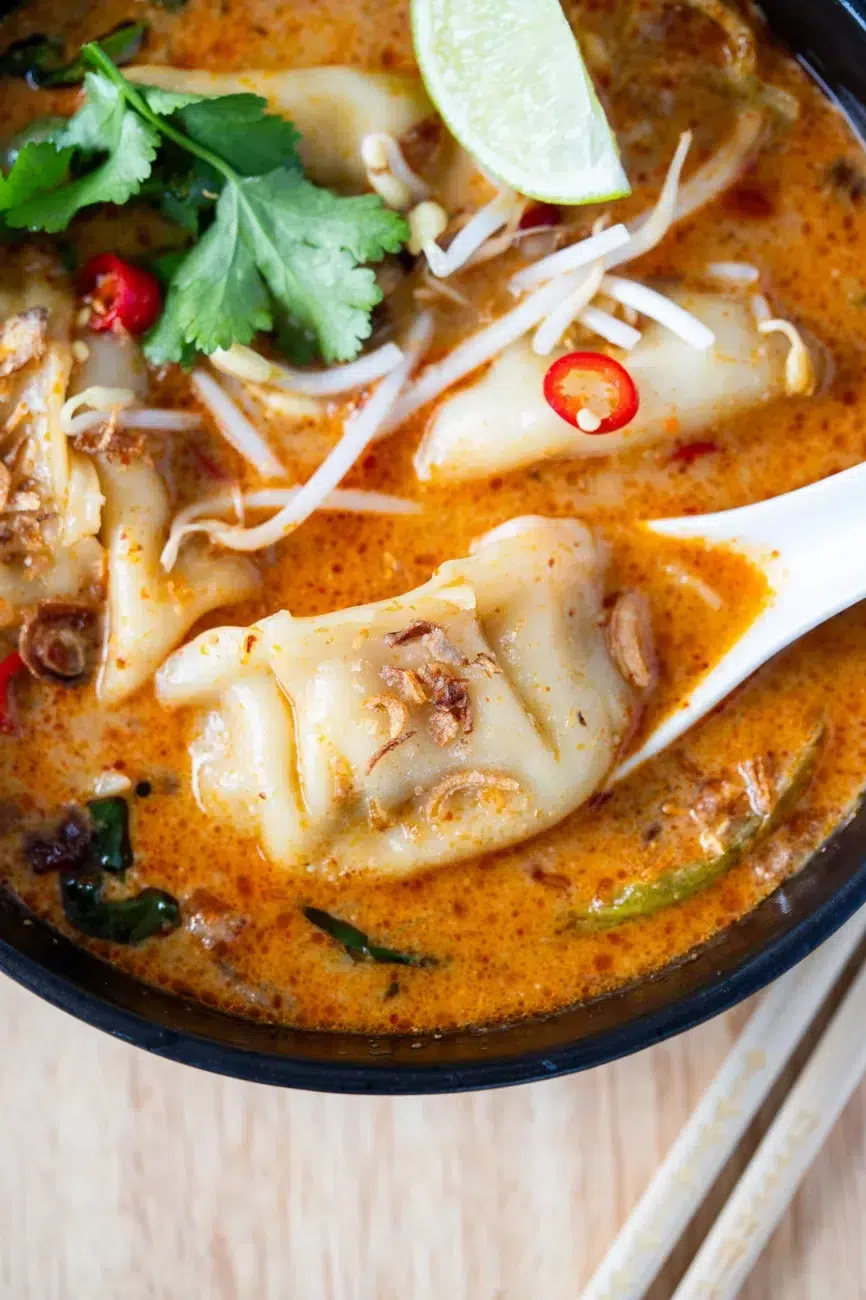 close up of creamy dumpling laksa soup in a black bowl and a lime wedge, with chopsticks resting on the side
