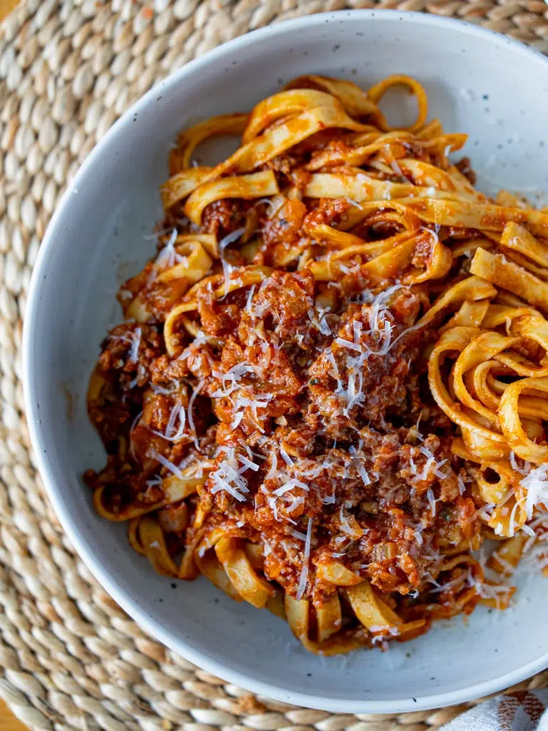 Top view of fettuccine pasta with hidden vegetable Bolognese sauce and grated Parmesan, served in a white speckled bowl on a woven placemat.